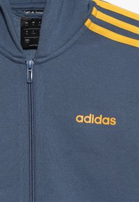 Blue zip-up jacket with yellow Adidas logo on the chest and three yellow stripes on the shoulder.