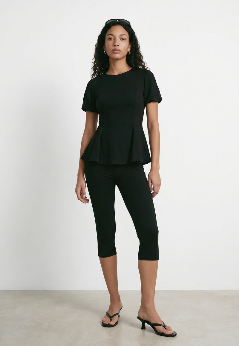 Femme portant un haut peplum noir à manches bouffantes, des leggings noirs courts, des sandales à talons noires et des lunettes de soleil sur la tête, debout devant un fond blanc.