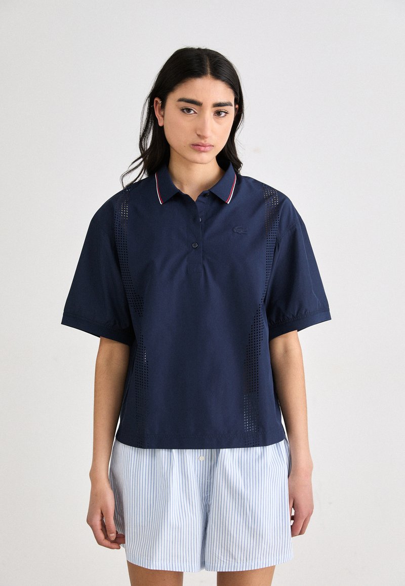 Marineblå polo-shirt med korte ærmer, med en knaplukning og en krave i rød og hvid stribe; mesh detalje på siden.