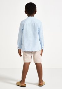 Ljusblå randig skjorta med knappar och långa ärmar, i kombination med beige shorts och bruna slip-on skor, sedd bakifrån.