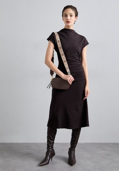 Brown handbag with a logo, paired with a sleeveless black dress, form-fitting with a high neckline, and brown knee-high boots with pointed toes.