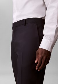 Άνδρας που φοράει μαύρο παντελόνι ραμμένο στα μέτρα του και λευκό πουκάμισο με μακριά μανίκια, με χαλαρό χέρι στο πλευρό, σε απλό φόντο.