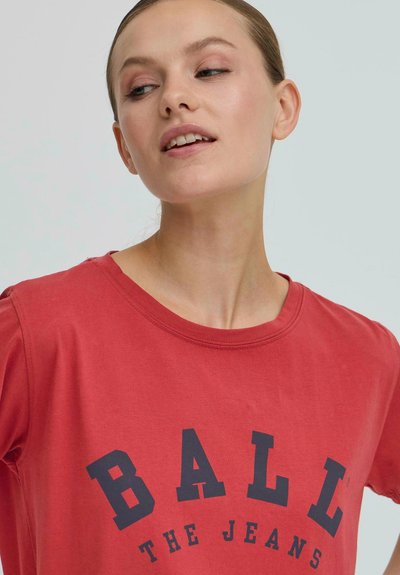 Jeune femme en t-shirt rouge avec le texte "BALL THE JEANS" regarde légèrement sur le côté devant un fond clair.