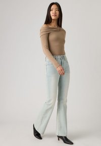 Νεαρή γυναίκα που φοράει μπεζ τοπ με γυμνούς ώμους, ανοιχτόχρωμα μπλε τζιν και μαύρα μποτάκια αστραγάλου, στέκεται μπροστά σε ένα απλό φωτεινό φόντο.