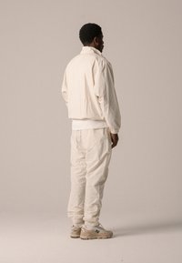 Veste blanche texturée avec un col haut, associée à un pantalon blanc ample et des baskets beiges, créant un look monochrome minimaliste.