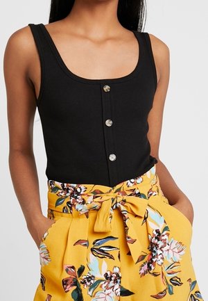 Zwarte tanktop van geribd stof met een ronde halslijn en een drieknoopsvesting aan de voorkant, gecombineerd met gele hooggetailleerde shorts met bloemenprint.