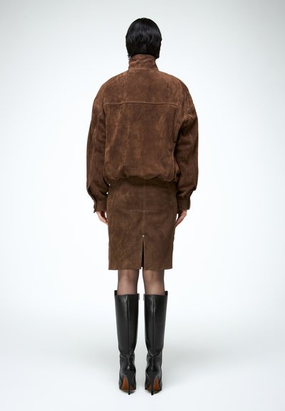 Persoon met kort donker haar, gekleed in een bruin suède jack en rok, zwarte laarzen met hak tot aan de knie, staand met de rug naar de kijker op een witte achtergrond.