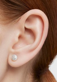 Primer plano de la oreja de una persona pelirroja que lleva un pequeño pendiente de plata redondo en forma de osito de peluche.