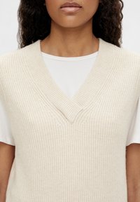 Femme portant un gilet sans manches à col en V tricoté de couleur crème, par-dessus une chemise blanche à manches courtes, avec des cheveux bouclés foncés visibles.