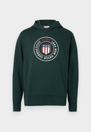 Dunkelgrüner Kapuzenpullover mit einem vorderen runden Grafikdesign, das ein Schild zeigt, sowie den Texten "GANT" und "1949 NEW HAVEN CONNECTICUT".