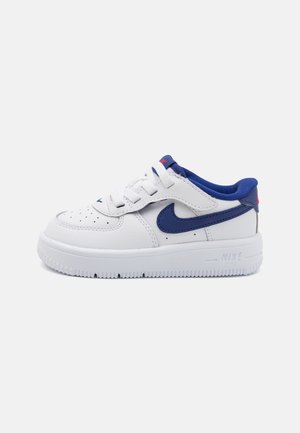 Hvid og blå Nike-sneaker med snørebånd og velcrolukning, med blå indvendig foring og sål med Nike-logo på siden.