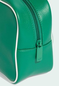 Zeleni toaletni neseser od teksturirane veganske kože s bijelim obrubom, s patentnim zatvaračem i zelenim privjeskom. Kompaktan i zaobljen oblik.