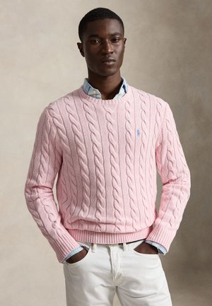 Mann trägt einen rosa Zopfpullover über einem blauen Hemd mit Kragen und weißen Hosen, Hände in den Taschen, steht vor einem neutralen Hintergrund.