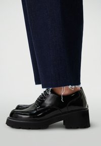 Chaussures à lacets en cuir verni noir avec une épaisse semelle noire. Elles ont un bout arrondi et des coutures décoratives, associées à un pantalon bleu foncé effiloché.