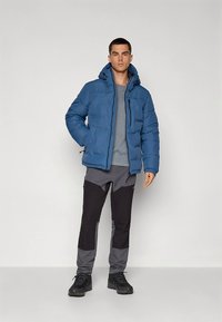 Zila pūkaina jaka ar kapuci, rāvējslēdzēju priekšpusē un elastīgiem aprocēm, kopā ar pelēkām un melnām krāsu blokā novietotām biksēm un melniem apaviem.