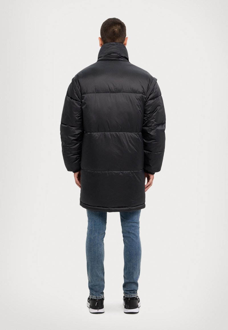 Abrigo acolchado negro con cuello alto, con un diseño acolchado y una silueta larga. Combinado con vaqueros azules y zapatillas negras.