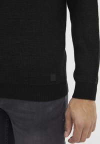 Strikket svart genser med teksturert stoff, ribbestrikkede mansjetter og kant. Har et lite svart merke på den nedre siden. Brukt over grå jeans.