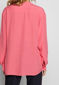 Woman wearing a loose-fitting, long-sleeve coral pink blouse with buttoned cuffs and black pants, shown from the back.