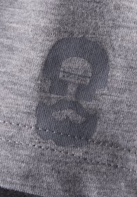 Grauer Stoff mit weicher Textur, versehen mit einem dunkelgrauen Druckmotiv eines stilisierten Gesichts mit einem Schnurrbart, das am unteren Rand platziert ist.