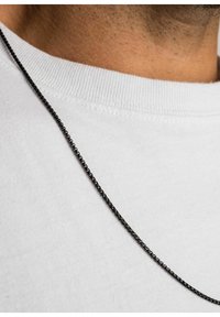 En sort metal kædedyse ligger på en hvid T-shirt med rund hals. Kæden har led i et fint, tekstureret mønster.