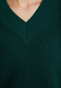 Gros plan sur une personne portant un pull côtelé vert foncé avec un col en V et des clavicules visibles.