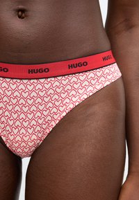 Červenobiely bikini so vzorom srdiečok a čiernym pásikom s nápisom "HUGO." Vyrobené z mäkkého materiálu, s nízkym pásom pre pohodlie.