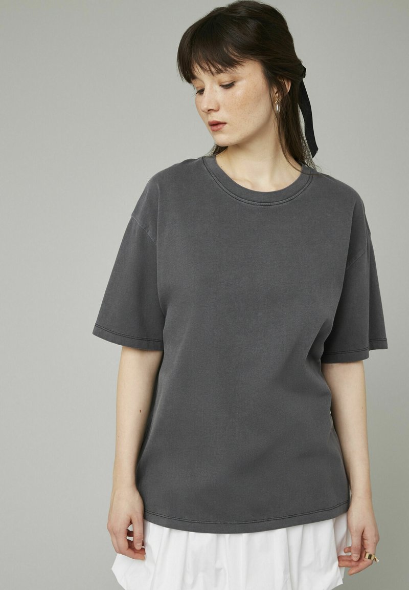 T-shirt gris à manches courtes en matière douce avec un col rond, coupe légèrement ample et détails de couture visibles. Assorti à une jupe blanche.