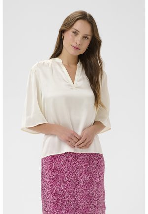 Femme aux longs cheveux bruns portant un chemisier blanc ample et une jupe rose à motifs, debout devant un fond clair uni.