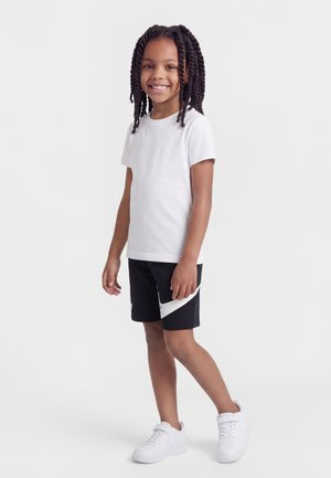 Χαμογελαστό παιδί με μακριά κοτσίδα, που φοράει λευκό μπλουζάκι, μαύρο-άσπρο σορτς και λευκά αθλητικά παπούτσια, στέκεται μπροστά σε ένα απλό γκρι φόντο.