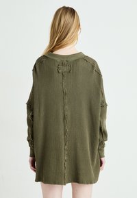 Olíva zöld, oversized pulóverruha texturált, gofris kötésű anyagból, lejjebb hasított vállakkal és nyers szélekkel az ujjakon és a hátsó varrásnál.