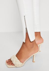 Pantalones blancos ajustados con detalle de cremallera plateada en el dobladillo, combinados con tacones de punta abierta beige trenzados sobre un fondo neutro.