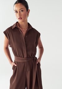 Bruine jumpsuit met een kraag, korte mouwen, knoopsluiting en een strik taille. Glad materiaal met een relaxte pasvorm.