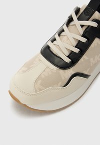 Beige sneaker with black glossy accents and light fabric. Features tonal logo pattern, flat laces, and a textured rubber sole.