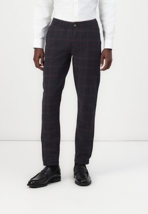 Mann trägt schwarz-rot karierte Hose, weißes Hemd und schwarze Lederloafers, steht vor einem einfachen weißen Hintergrund.