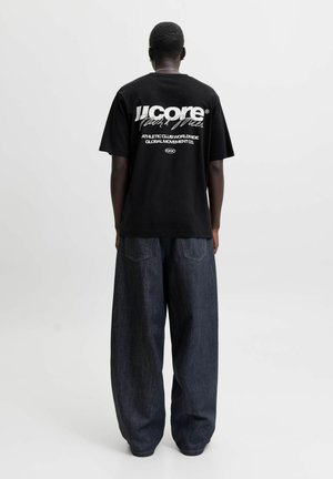 Homme portant un t-shirt graphique noir et un jean bleu foncé ample, debout de dos sur un fond uni.