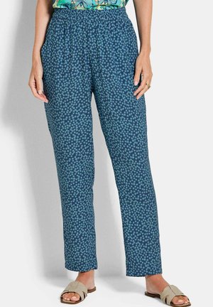 Donna che indossa pantaloni larghi blu con un motivo floreale azzurro chiaro e sandali beige aperti in piedi davanti a uno sfondo semplice.