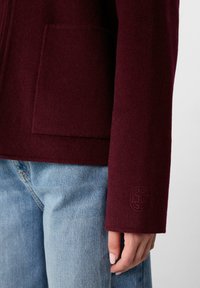 Primo piano di una mano vicino alla tasca di un cappotto borgogna indossato con jeans azzurri, che mostra il logo ricamato sulla manica del cappotto.