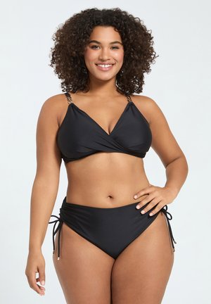 Lachende vrouw met krullend haar die een zwarte bikini draagt met kettingbandjes en zijstrikjes aan de broek, staand tegen een lichte achtergrond.