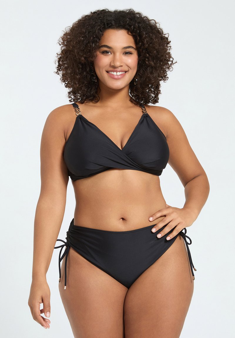 Mujer sonriente con cabello rizado que lleva un bikini negro con tirantes de cadena y braguitas con lazos laterales, de pie frente a un fondo claro.