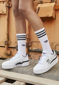 White leather sneakers with black accents, textured sole, and perforated toe cap. Paired with white socks featuring black stripes and logo.