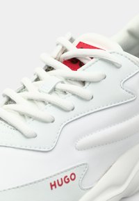 White sneaker with textured upper, white laces, and red "HUGO" logo. Features padded collar and tongue, smooth soles with curvy design.