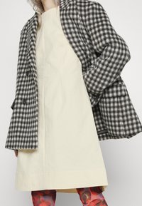 Abrigo de lana gris y blanco con cuadros sobre un vestido crema; tejido texturizado; mallas de patrón colorido visibles en la parte inferior.