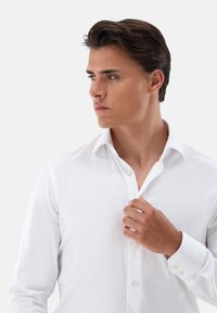 Camicia bianca con bottoni, collo classico, tessuto liscio e silhouette aderente. Presenta bottoni visibili e una parte superiore leggermente sbottonata.