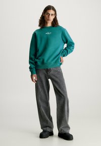 Felpa teal con colletto e polsini a coste, abbinata a jeans grigi larghi. Il modello è in piedi con le mani sui fianchi, indossando scarpe nere.