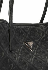 Sac à main noir matelassé en cuir texturé, avec un logo Guess en or triangle sur le devant, équipé de poignées doubles.