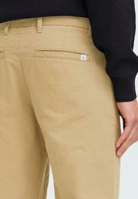 Pantalons en coton beige avec une texture lisse, dotés de deux poches arrière et d'une petite étiquette de logo. La taille est plate avec une coupe standard.