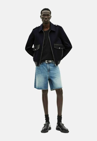 Giacca navy con dettagli in pelle nera, camicia nera, pantaloni corti in denim chiaro e scarpe nere. Il modello ha i capelli corti e si trova in piedi con le mani nelle tasche.