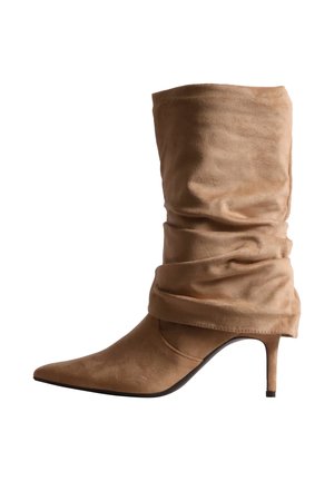 Botte en daim beige fauve montant jusqu’au genou, avec une tige souple et plissée, un bout pointu et un talon haut et fin, sur fond blanc.
