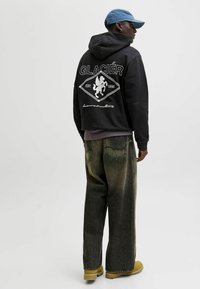 Sudadera negra con gráfico blanco en la espalda, con la inscripción "GLACIER EST. 1990". Combinada con jeans de mezclilla de pierna ancha y desgastados, y botas color miel.