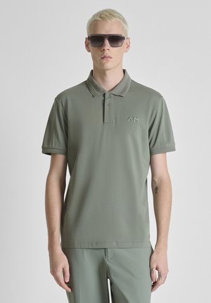 Blonde man met donkere zonnebril, groene poloshirt met AM-logo en bijpassende groene broek, staand tegen een effen lichte achtergrond.